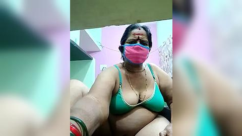 Media: Video of a middle-aged South Asian woman with medium skin tone, wearing a turquoise bra, pink face mask, and a beaded necklace. She's in a dimly lit room with pink walls and a green cabinet.