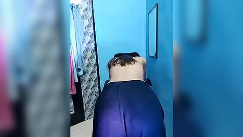 Media: Video of a person with a pale skin tone and dark hair in a ponytail, wearing a black sports bra and black leggings, doing yoga in a room with blue walls and floral-patterned curtains.