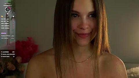 Media: Video of a smiling, light-skinned young woman with long, straight brown hair, wearing a delicate gold necklace, in a dimly-lit room with blurred background.