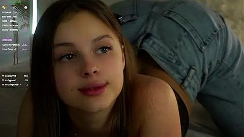 Media: A video of a young Caucasian woman with straight brown hair and light skin, wearing a black tank top, lying on a bed with blue jeans partially covering her.