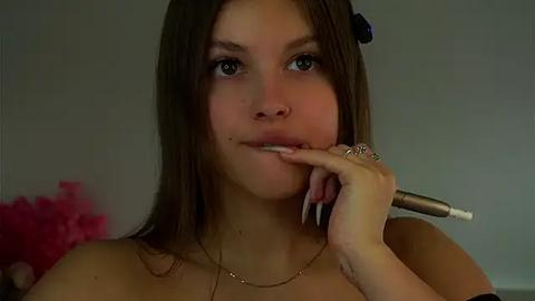 Media: Video of a young woman with fair skin, brown hair, and blue eyes, holding a mascara wand near her lips, wearing a gold necklace.