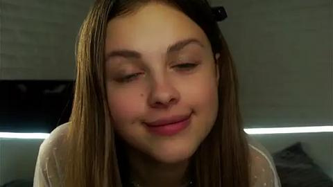 Media: Video of a young Caucasian woman with long brown hair, wearing a black hair clip, smiling gently, in a dimly lit room.