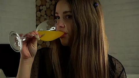 Media: Video of a young woman with long brown hair, wearing a black sheer top, drinking orange juice from a wine glass, in a modern, dimly lit room with a wood pile in the background.