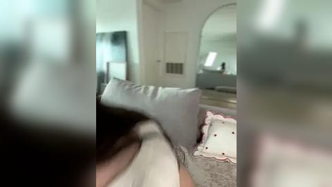 Media: A video of a blurry, partially obscured bedroom featuring a bed with white and pink linens, a mirror, and a door in the background.