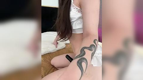 Media: A video of a slender, fair-skinned woman with long brown hair, wearing a white lace lingerie set, sitting on a bed with a brown comforter. Her right thigh is adorned with a black tribal tattoo. The room has black and white walls.