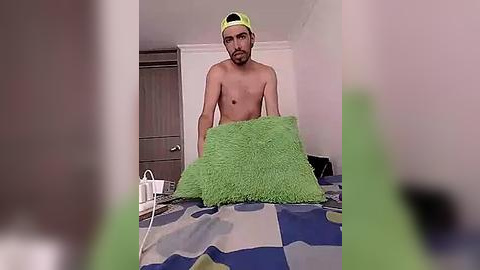 Media: Video of a shirtless, bearded man in a green baseball cap, holding a large green pillow in a room with a bed covered in a blue and green patterned blanket.