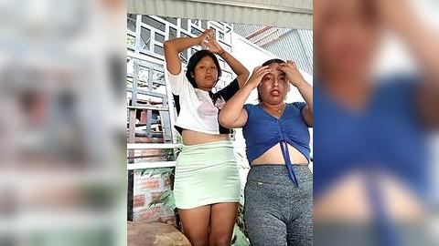 Media: Video of two women with medium builds and short black hair, wearing casual outfits: one in a white crop top and light green skirt, the other in a blue tie-dye top and grey pants. Background shows a partially blurred outdoor scene with a metal staircase.