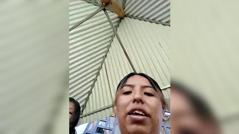 Media: A video showing a man with a medium skin tone, short black hair, and a neutral expression, holding a beige umbrella with a corrugated metal roof. The background features blurred people and a concrete structure.