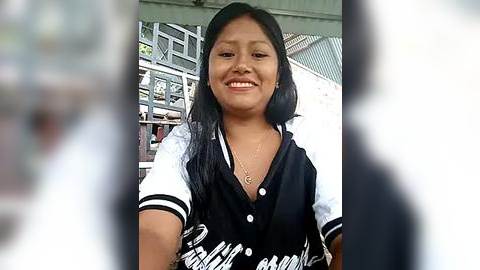 Media: Video of a smiling young woman with medium brown skin and straight black hair, wearing a black and white varsity jacket, in front of a blurred market scene.