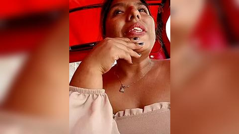 Media: Video of a Latina woman with dark hair, medium brown skin, and full lips, wearing a beige off-shoulder top, eating a chocolate bar, with a blurred red background.