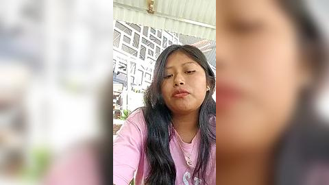 Media: Video of a young Asian girl with long black hair, wearing a pink shirt, sitting outdoors under a corrugated metal roof. Background shows blurred structures and greenery.