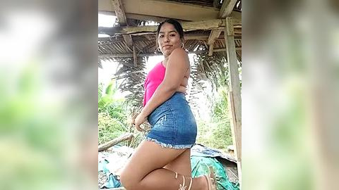 Media: A video of a Latina woman with medium skin tone, wearing a pink tank top and frayed denim shorts, kneeling in a rustic outdoor setting with thatched roof and palm trees in the background.