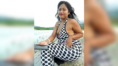 Media: Video of a smiling East Asian woman in a black-and-white polka dot halter dress, sitting on a rocky shore with a calm lake in the background.