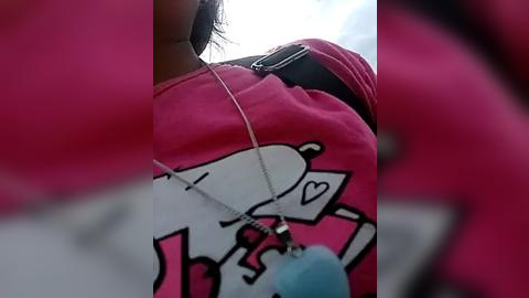 Media: Video of a woman wearing a pink T-shirt with a cartoon dog graphic. She's holding a blue plush toy and a black phone. Background is blurred.