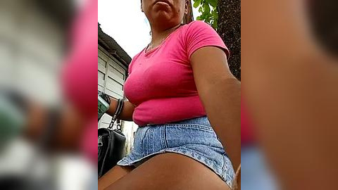 Media: Video of a woman in a tight pink top and high-waisted denim shorts, carrying a black handbag, outdoors with blurred background.