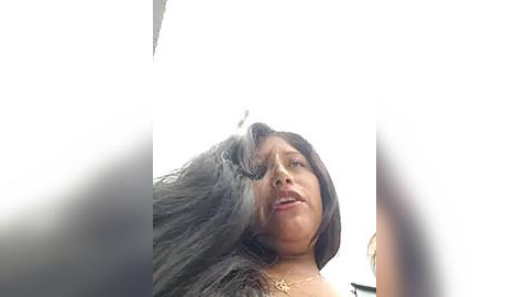 Media: Video of a woman with long, straight black hair, light brown skin, and a somber expression, taken from an unusual, slightly blurred perspective.