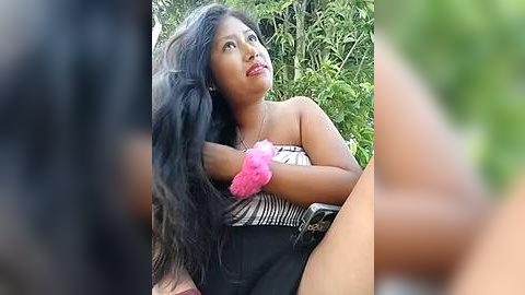 Media: Video of a young Southeast Asian woman with long, black hair, wearing a pink scrunchie, black shorts, and a striped top, sitting outdoors with lush green foliage in the background.