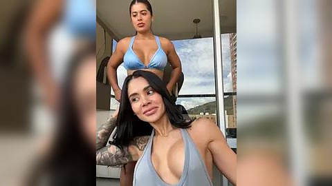 Media: Video: Two women, one with a blue bikini top, another with tattoos and a gray tank top, in a modern living room with large windows and a cityscape view.