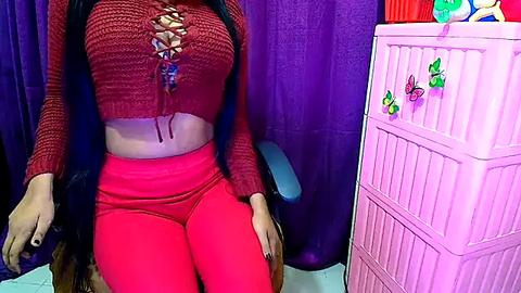 Media: Video of a woman with a medium complexion wearing a red knitted crop top and matching high-waisted leggings, sitting on a purple chair. The background features a pink baby crib with colorful butterfly decals and purple curtains.