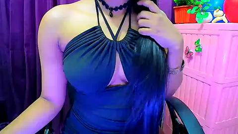 Media: Video of a light-skinned woman with a curvy figure, wearing a teal halter dress with a deep cutout, black choker, and holding a phone. Background includes a pink wall with decor, purple curtain, and a plant.