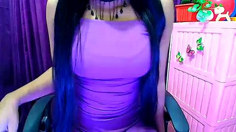 Media: Video of a woman with long, dark blue hair wearing a tight, light purple dress with a ruched bodice, seated in a chair. Background features a pink crib with colorful decals and a purple curtain.