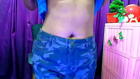 Media: Video of a light-skinned person with a flat stomach wearing blue camouflage shorts, standing against purple curtains. Background includes a white dresser with green plants and a red object, possibly a plush toy.