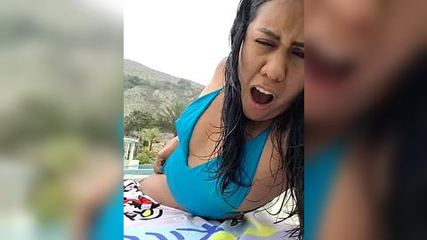 Media: Video of a woman with long, wet black hair in a turquoise bikini, screaming in pain, lying on a colorful towel by a pool, with a blurred man in the foreground.