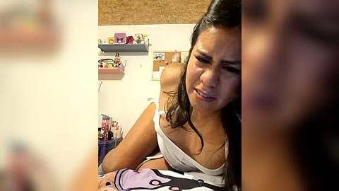 Media: Video of a young woman with dark hair, wearing a white tank top, sitting on a bed, looking distressed, surrounded by cluttered shelves and personal items in a small, messy room.