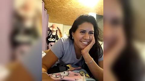 Media: Video of a smiling woman with long black hair, wearing a light blue t-shirt, lying on a bed with a cartoon-patterned blanket in a cozy bedroom with wooden ceiling.