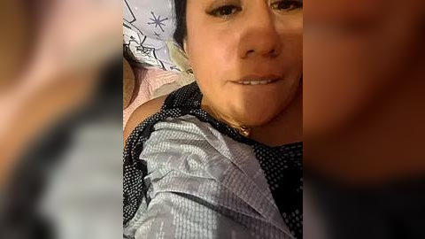 Media: Video of a young woman with light skin, dark hair, and a slight smile, wearing a gray and black patterned shirt. She's lying on a white pillow with a snowflake design, in a blurred background.