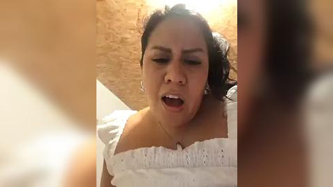 Media: Video of a Latina woman with dark hair, wearing a white blouse, screaming in pain, set against a brick wall.