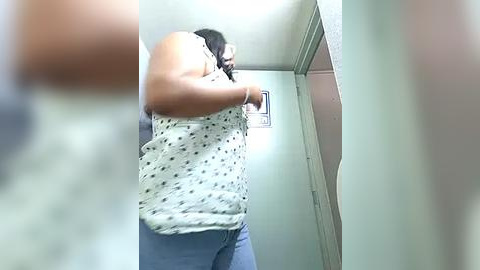 Media: Video of a woman with medium skin tone, wearing a white sleeveless top with small black polka dots and blue jeans, taking a selfie in a narrow, dimly lit hallway.