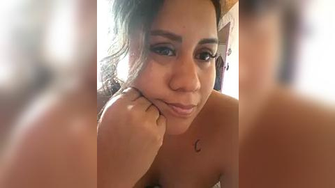 Media: A close-up video of a young Latina woman with medium brown skin, dark hair, and full lips, smiling slightly, leaning on her hand, with a crescent moon tattoo on her chest.