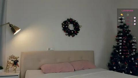 Media: Video of a modern bedroom with a beige upholstered headboard, two pink pillows, a black wreath on the wall, a silver lamp on the left, and a lit Christmas tree on the right.