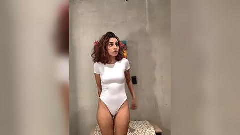 Media: Video of a young woman with curly brown hair, wearing a white bodysuit, standing in a minimalist room with concrete walls and abstract art.