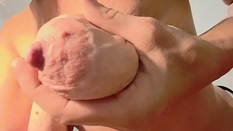 Media: A video shows a close-up of a nude, fair-skinned woman with large, round breasts. She is cupping and squeezing her right breast with both hands, highlighting the nipple and areola. The background is blurry, with a soft, warm light illuminating her skin.