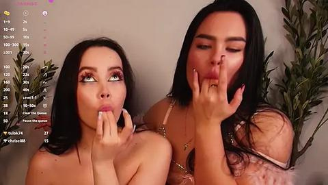 Media: Video of two women with long black hair, one with green eyes and the other with brown eyes, kissing each other's fingers. They're in a room with a calendar and potted plants in the background.