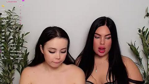 Media: A video of two women with long black hair, wearing makeup, against a white background. One woman is topless, the other in a black strapless top.