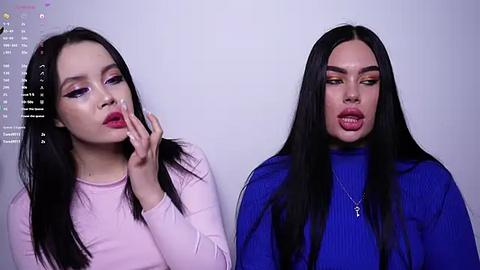 Media: Video of two young women with long black hair, one in a pink top, the other in a blue top, both with exaggerated makeup, against a plain white background.