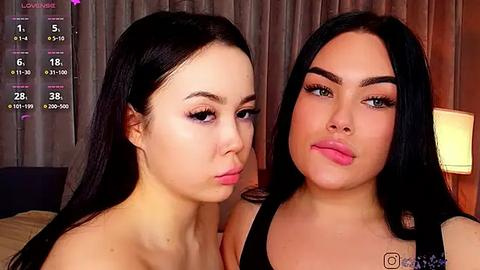 Media: Video of two young women with long black hair, fair skin, and natural makeup; one puckers her lips, the other has a neutral expression. Background shows a dimly lit bedroom with a bed and curtains.