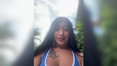 Media: Video of a young woman with long black hair, medium skin tone, wearing a blue bikini top, and a tattoo on her left shoulder. Background is blurred, showing greenery.