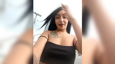 Media: A video of a Latina woman with long black hair, medium skin tone, and a tattoo on her right shoulder. She wears a black, off-the-shoulder crop top, smiling and covering her eyes with her hand. Background is blurred.