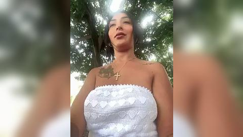 Media: Video of a woman with medium skin tone, wearing a white, lace-trimmed strapless top, standing outdoors among green foliage.