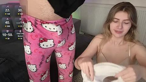 Media: Video of a young woman in pink Hello Kitty pajamas, sitting on a bed, watching a man in black shorts pull down his pants.