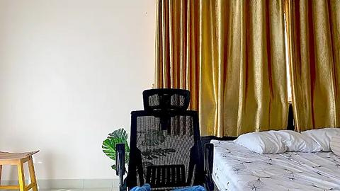 Media: A video of a minimalist bedroom with a black mesh office chair in the center, facing a bed with white sheets and a golden curtain backdrop.