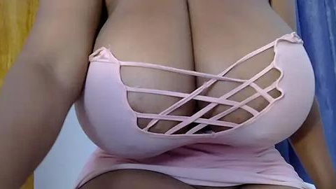 Media: Video of a woman with large, cleavage-revealing, pink halter top featuring crisscross straps, revealing ample cleavage. Background features yellow and blue curtains.