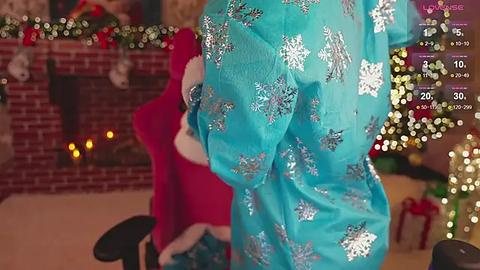 Media: Video of a person in a turquoise onesie with white snowflake patterns, standing next to a red Santa suit, in a cozy room with a lit Christmas tree, fireplace, and festive decorations.