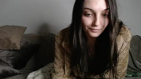 Media: Video of a young woman with long, straight dark hair, wearing a beige fuzzy sweater, sitting on a bed with gray sheets and pillows, looking contemplative.