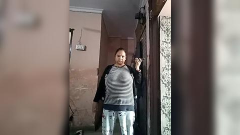 Media: Video of a middle-aged, heavyset woman with short hair, wearing a striped shirt and patterned pants, standing in a narrow hallway with peeling beige walls, patterned wallpaper, and a partially open door.