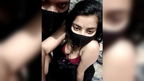 Media: Video of a young woman with long black hair, wearing a black mask, maroon top, and black jacket, sitting on a bed, surrounded by blurred figures.